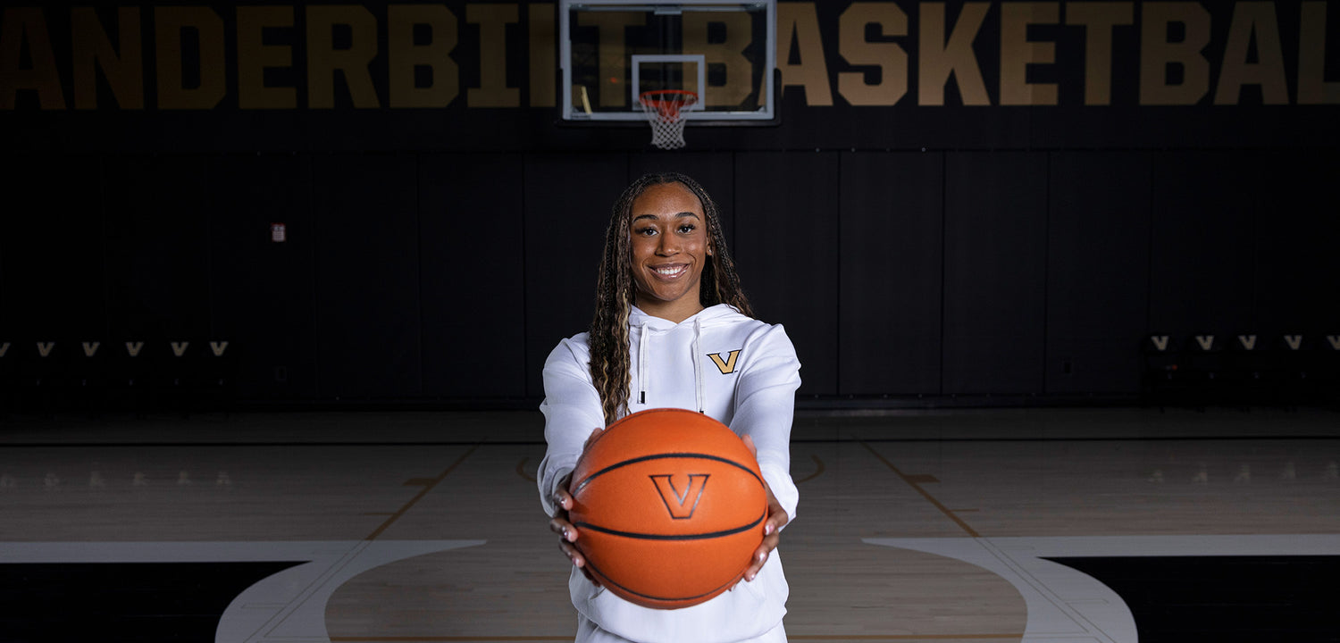 Vanderbilt women’s basketball player standing on an indoor court, smiling and holding a basketball toward the camera with a hoop and ‘Vanderbilt Basketball’ signage in the background.