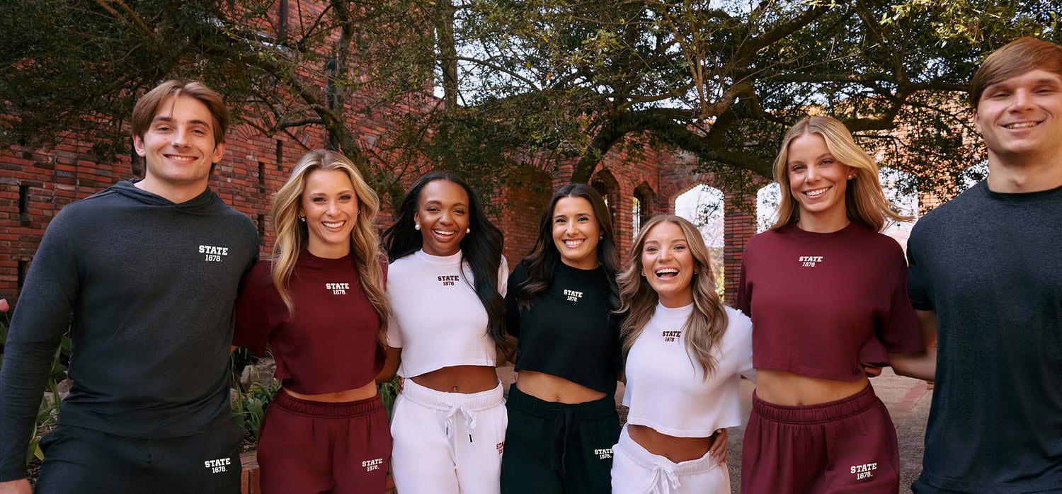 Group of Mississippi State students wearing maroon, white, and black apparel, posing outdoors in front of a brick campus walkway with trees.