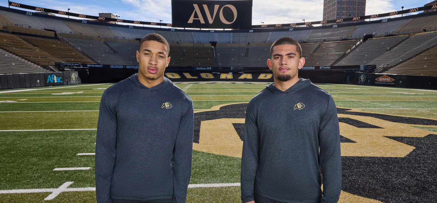 Two male athletes wearing dark Colorado Buffaloes hoodies stand side by side on a football field inside a large stadium, with an “AVO” sign displayed on the scoreboard behind them.